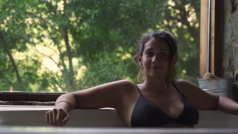 Young-attractive-female-having-a-fancy-bath-in-a-jacuzzi-with-big-green-nature-window-looking-to-camera-smile-able-to-slow-motion-60fps
