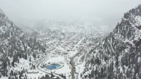 Aerial-view-of-Ouray,-Colorado-USA,-valley-town-and-hot-geothermal-spring-water-pools-in-white-foggy-snowy-winter-landscape,-drone-shot