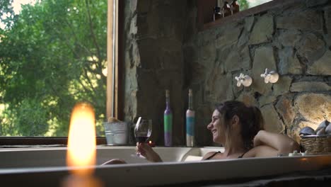 Young-attractive-female-having-a-fancy-bath-in-a-jacuzzi-drinking-wine-looking-by-big-green-nature-window-able-to-slow-motion-60fps