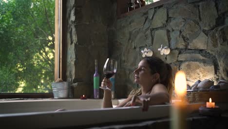 Young-attractive-female-having-a-fancy-bath-in-a-jacuzzi-drinking-wine-with-big-green-nature-window-able-to-slow-motion-60fps
