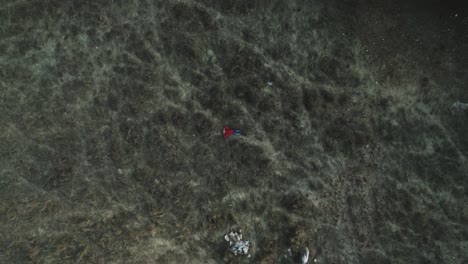 Top-down-aerial-view-of-relaxed-young-man-wearing-red-sweater-and-jeans-looks-up-sky-lying-on-dark-grass-field,-drone-rising-reveal-forest-by-road,-day