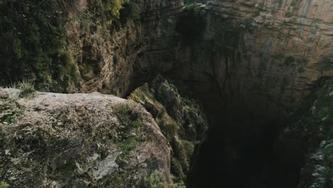 Sensational-aerial-view-of-arch-rock-formation-in-Lebanon-mountain-landscape,-young-hiker-man-wearing-sporty-clothes-leans-over-cliff,-drone-flying-backwards,-day