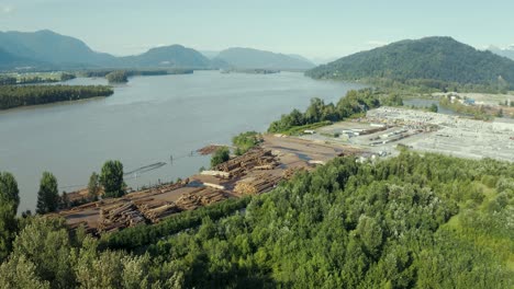 Cinematic-drone-shot-of-a-tree-logging-processing-plant-along-a-river-bank-surrounded-by-mountains