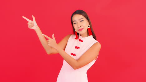 Joven-Mujer-Asiática-Sonriente-Apuntando-A-La-Izquierda-Con-Los-Dedos,-Aislada-En-Un-Fondo-Rojo-Para-Adiciones-Interactivas,-Logotipo-O-Eslogan,-Fotograma-Completo