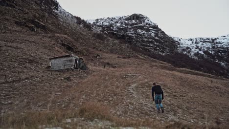 Young-man-hiking-through-abandoned-meadow-with-small-wooden-shed-and-snow-covered-mountains-in-the-background