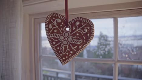 Red-gingerbread-heart-hanging-in-a-window,-snow-covered-trees-in-background