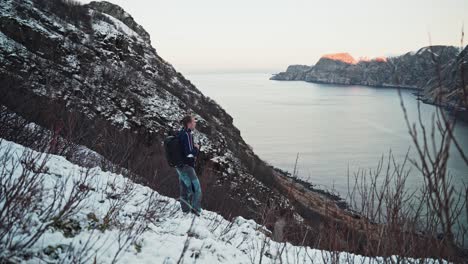 Young-man,-walking-on-the-edge-of-a-snow-covered-mountain,-standing-still-to-take-in-the-scenery,-looking-over-a-beautiful-mountain-and-ocean-view