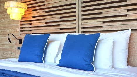Tilt-down-from-a-modern-wood-slat-headboard-to-a-hotel-bed-covered-with-bed-pillows-and-decorative-blue-pillows-and-blanket