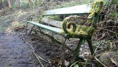 Mossy-neglected-weathered-green-curled-wrought-iron-wooden-bench-abandoned-in-woodland-forest