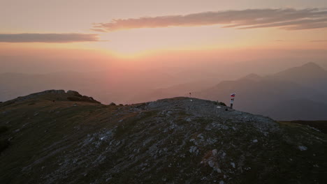 Drone-shot-of-the-top-of-the-mountain-at-the-sunrise,-hiker-holding-a-slovenian-flag-on-the-pol-which-is-fluttering-in-the-breaze