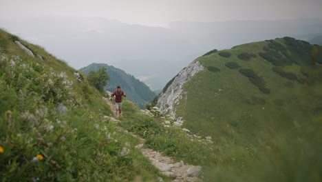 A-young-hiker-waliking-on-a-narrow-path-up-a-hill-with-hiking-poles-an-carrying-a-grey-backpack-in-background-are-visible-some-other-mountains-but-are-covered-in-low-cloud-cover