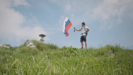 Camera-tracking-from-low-perspective-up-a-hill-a-hiker-at-the-top-with-a-hiking-pole-in-his-hand-and-a-slovenian-flag-attached-on-it-and-letting-it-flutter-in-the-wind