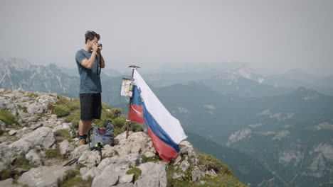 Excursionista-Parado-En-La-Cima-De-La-Montaña-Raduha,-Parado-Junto-A-La-Bandera-Eslovena-Y-Su-Mochila-Tomando-Una-Foto-Con-Una-Cámara