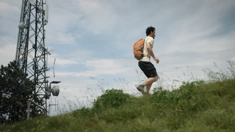 Camera-tracking-a-hiker-with-an-orange-backpack-running-up-a-hill-past-the-radio-tower-on-a-rocky-path-surrounded-with-green-summer-grass