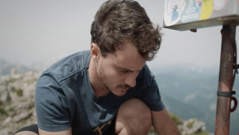 Young-hiker-squatting-at-the-top-of-mountain-by-a-box-to-stamp-his-hiking-booklet