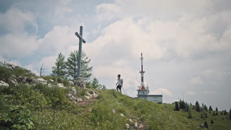 Camera-tracking-backwards,-hiker-walking-away-from-a-radio-tower-on-mountain-Uršlja-gora,-walking-past-a-big-cross