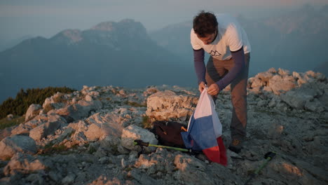 On-top-of-mountain-Peca-a-young-hiker-taking-a-slovenina-flag-out-of-his-backpack-in-the-earl-morning