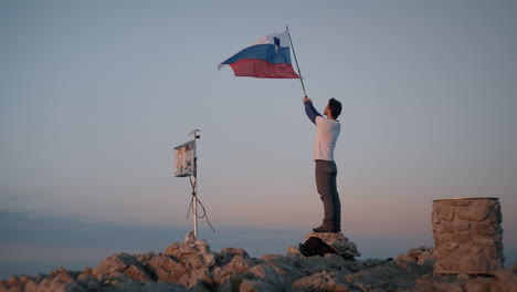 Man-standing-on-a-rock-and-holding-a-hiking-pole-in-the-air-with-attaches-slovenian-flag