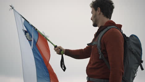 A-young-hiker-in-red-jacket-and-with-grey-backpack-holding-a-hiking-pole-on-which-he-attached-a-slovenian-flag-and-it-flutters-in-the-wind