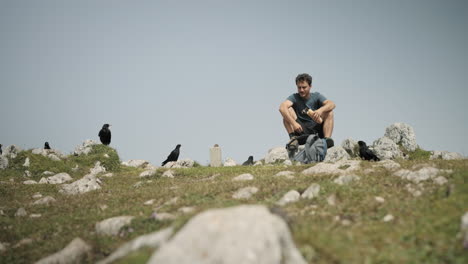 Hiker-sitting-on-the-rocks-and-eating-a-sandwich,-surrounded-by-alpine-choughs-waiting-for-a-small-share-of-food