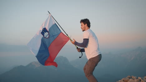 Man-holding-a-hiking-pole-on-which-he-attached-a-slovenian-flag,-steps-on-the-rock-and-lets-the-flag-flutter-in-the-wind