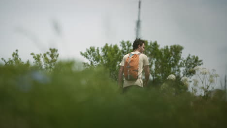 Hiker-with-an-orange-backpack-walkig-toweards-the-radio-tower-on-mountain-Slavnik,-camera-behind-greenery-on-low-perspective