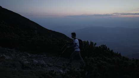 Camera-tracking-hiker-from-the-side-who-is-walking-with-hiking-polesa-up-a-hill-in-the-dawn-on-rocky-path-surrounded-with-conifers