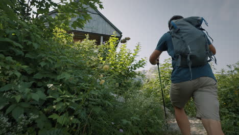 Camera-tracking-a-young-hiker-walking-up-a-path-towards-the-wooden-cottage-surrounden-with-green-plants