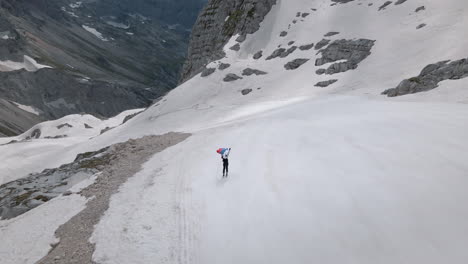 Tiro-De-Dron-De-La-Montaña-Kanin,-Un-Esquiador-Sosteniendo-Una-Bandera-Eslovena-Sobre-Su-Cabeza-Y-Esquiando-Cuesta-Abajo-Lejos-De-Un-Hombre