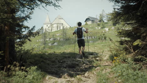 Hiker-walking-up-a-hill,-church-and-a-house-at-the-top,-thin-clouds-on-the-sky
