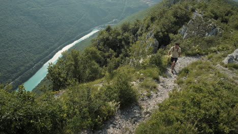 Hiker-with-a-backpack-walking-on-a-rocky-path-on-mountain-Sabotin,-top-view,-great-view-of-a-valley-with-a-torquoise-river-and-green-forests