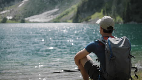 Hiker-sitting-by-the-mountain-lake-with-the-hiking-poles-and-a-backpack,-mountains-in-the-back,-water-is-glistening