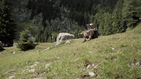 Camera-tracking-forward-towards-the-hiker-who-is-sitting-on-the-ground-on-green-grass-with-hiking-poles-in-his-hands