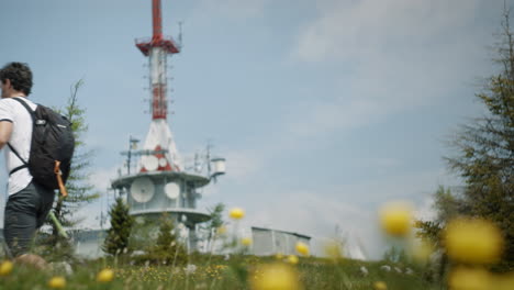 Hiker-walking-with-hiking-poles-past-the-camera-in-foreground-are-yellow-flowers-and-in-the-backgroun-is-radio-tower