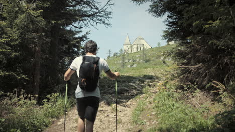 Hiker-climbing-up-a-hill,-walking-with-hiking-poles-past-the-conifers-and-towards-the-church-which-is-standing-at-the-top-of-mountain