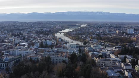 Aerial-flying-over-Kutaisi-city-with-snowy-mountains-and-river