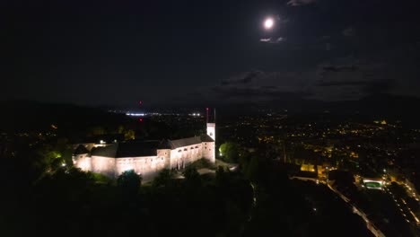 Night-aerial-hyperlapse-of-Ljubljana-castle,-flying-under-full-moon-moonlight,-city-lights-drone