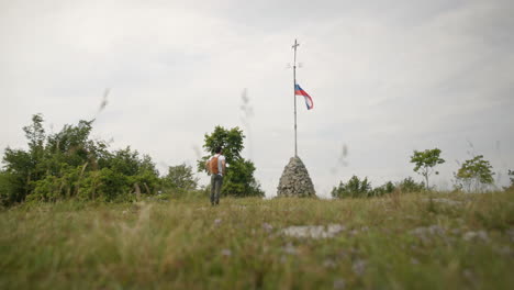 Hiker-with-a-backpack-walking-towards-the-rock-structure-with-a-slovenian-flag-and-a-cross-at-the-top-of-a-pole