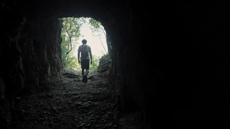 Hiker-with-an-orange-backpack-in-summer-clothes-walking-throug-a-dark-tunnel-carved-in-the-rock-towards-the-light-where-are-visible-green-trees