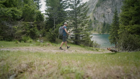 Hiker-with-the-hat,-backpack-and-hiking-poles-walking-towards-the-mountain-lake,-surrounded-by-conifers,-mountain-the-back