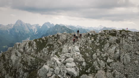 Hikers-standing-on-the-rocks-at-the-top-of-mountain-Rombon,-beautiful-moutanins-seen-in-background,-slovenian-flag-flutters-at-the-top