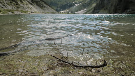 A-shot-of-the-edge-of-lake,-water-waving,-branch-in-the-water-in-background-is-valley-and-mountains