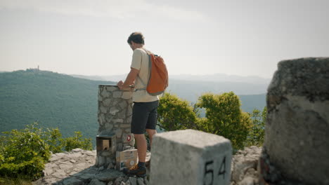 Hiker-with-an-orange-backpack-on-the-top-of-mountain-Saboti-standing-by-a-rock-monumet-with-a-compass-plate-for-direction-of-cities-or-places