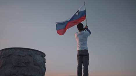 Camera-circling-aroud-the-hiker-at-the-top-of-mountain-Peca-holding-a-pol-with-a-slovenian-flag-attached-on-it,-fluttering-in-the-wind