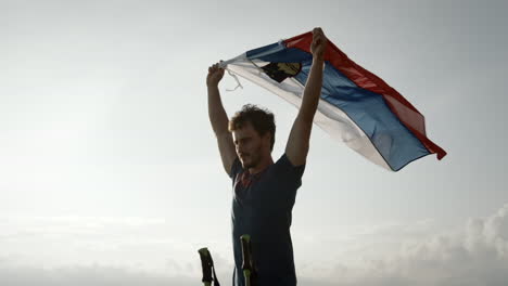 A-young-hiker-holding-in-his-hands-a-slovenian-flag-above-his-head-and-let-it-flutter-in-the-wind,-camera-cicling-arond-him