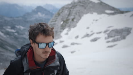 Hiker-climbing-on-mountain-Kanin-wearing-sunglasses,-mountains-parially-covered-in-snow-in-background