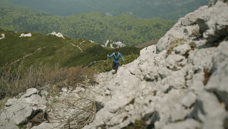 Hiker-climbing-on-a-rocky-path-towards-the-peak-of-mountain-Snežnik,-helping-himself-with-hiking-poles-with-balance
