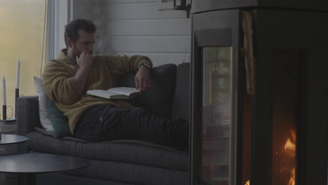 Caucasian-man-sits-in-sofa-reading-book-with-warm-fireplace-in-the-foreground,-rainy-cold-autumn-outside