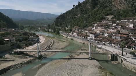 Aerial-shot-of-people-moving-over-bridge-and-ancient-houses-besides-river-in-Berat,-Albania