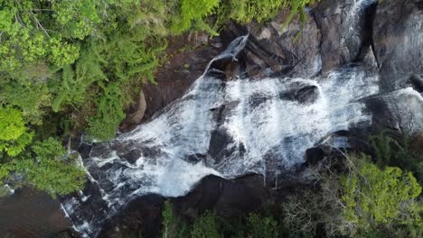 Drone-with-camera-down-making-slow-upward-movement-of-a-beautiful-waterfall,-flowing-waters-and-exuberant-nature-around
